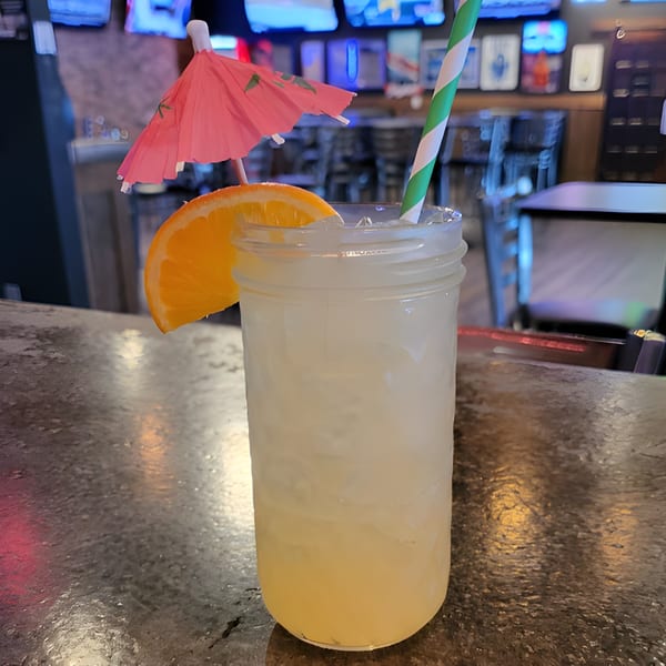 A refreshing cocktail with an orange slice, pink umbrella, and striped straw on a bar.