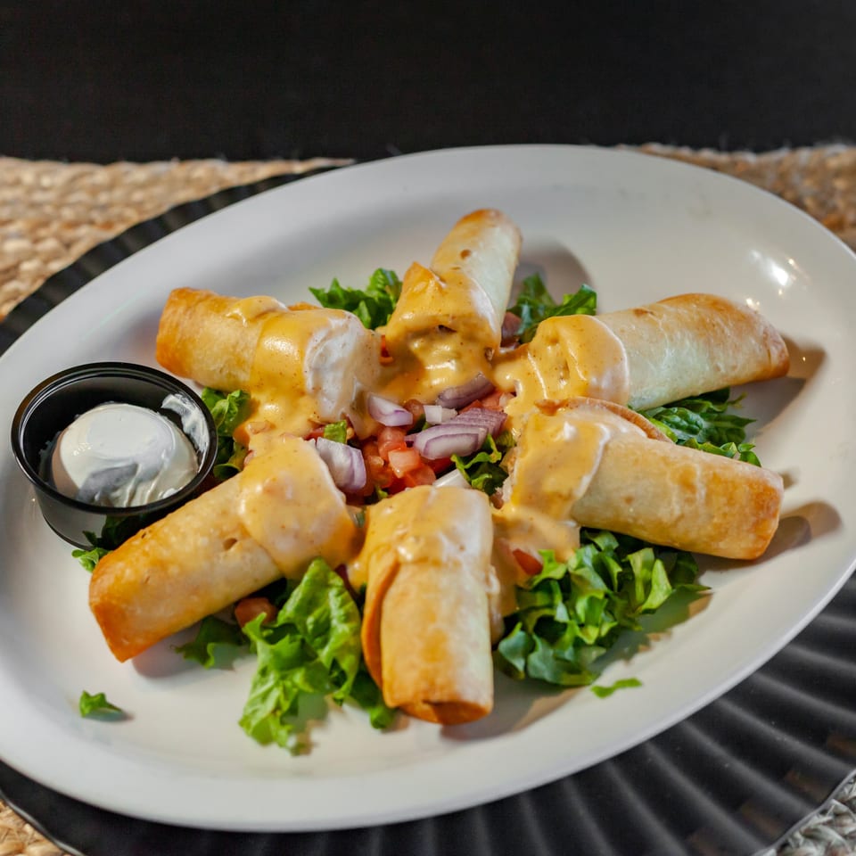 Taquitos with creamy sauce, pico de gallo, and a side of sour cream on a bed of lettuce.