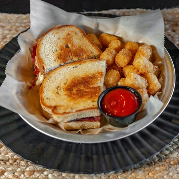 A grilled sandwich and tater tots with a side of ketchup in a metal basket.