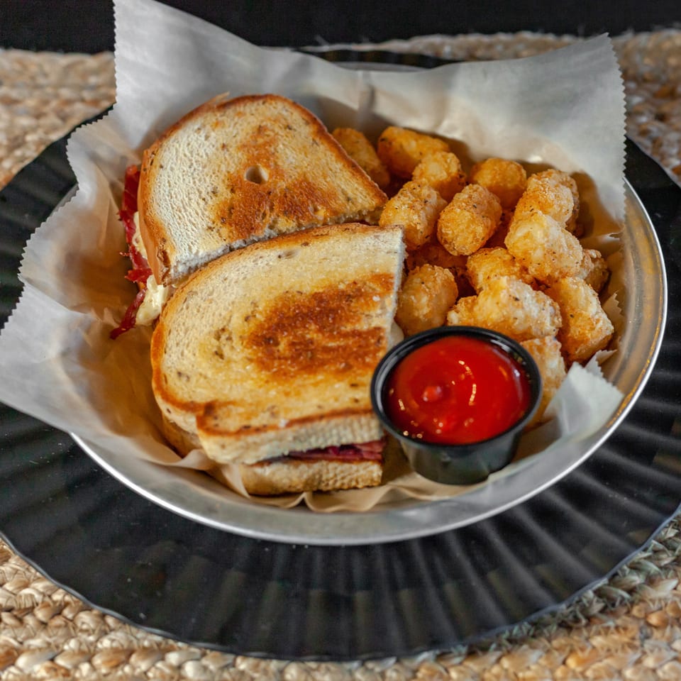 A grilled sandwich and tater tots with a side of ketchup in a metal basket.