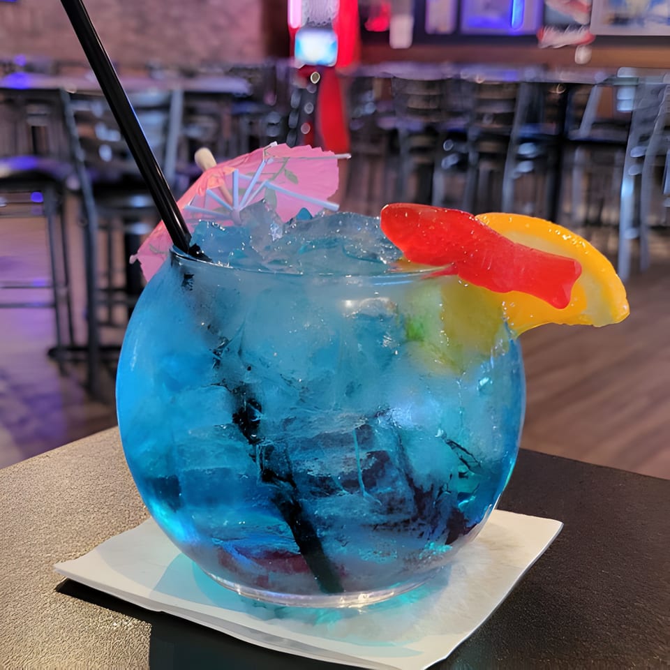 A vibrant blue cocktail with ice, a pink umbrella, and gummy candy in a sports bar setting.