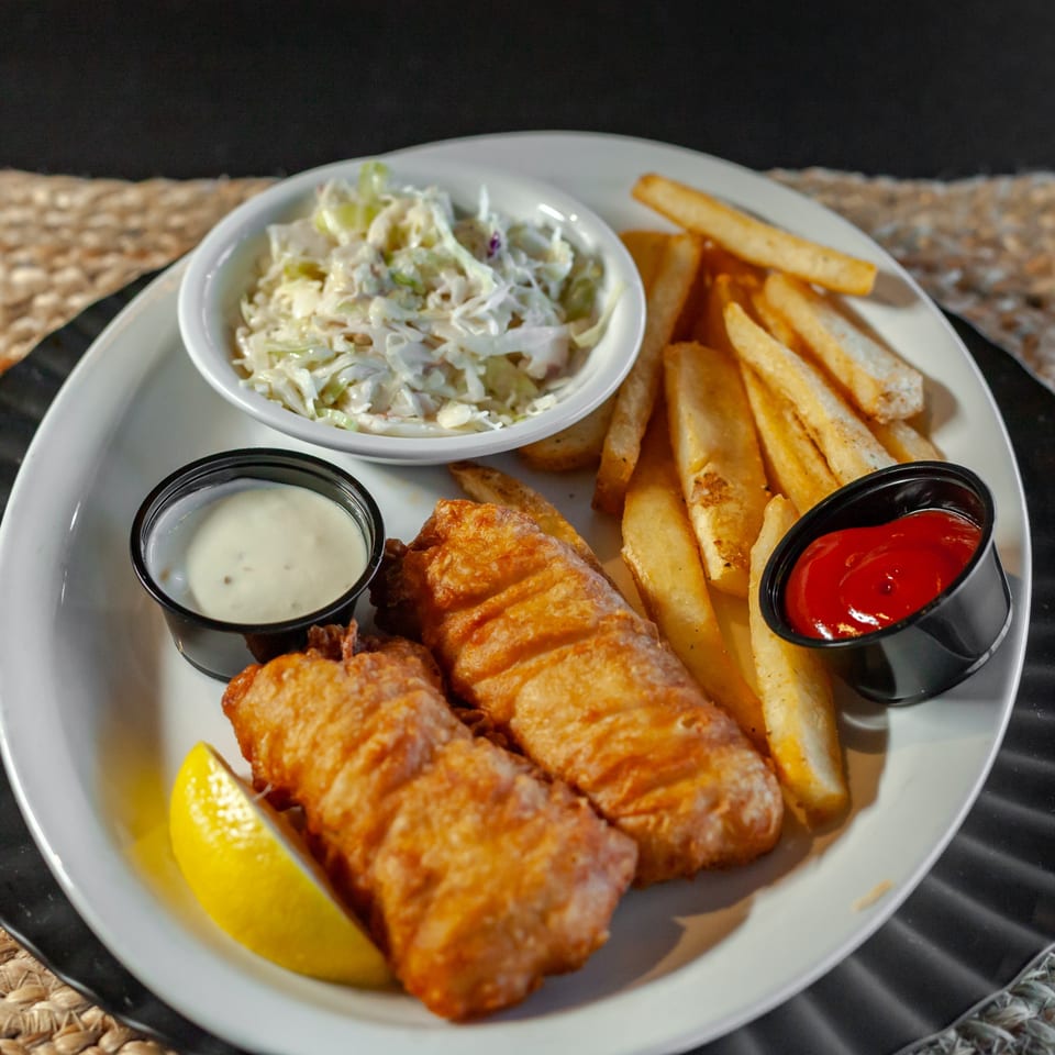 Golden-brown fish and chips with coleslaw, tartar sauce, ketchup, and a lemon wedge on a white plate.