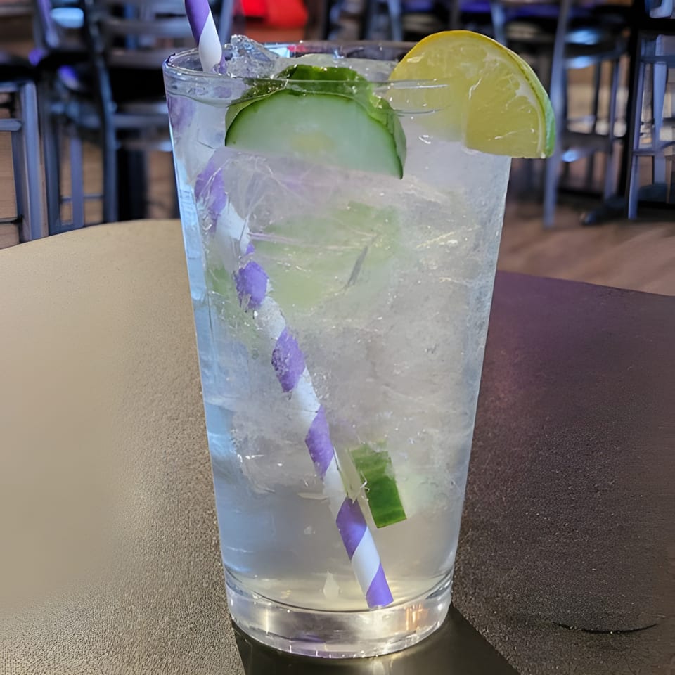 A refreshing drink with cucumber and lime slices, a purple and white straw, on a dark table.